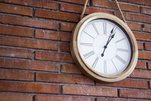 Medium Close Shot Of A Wooden Classic And Simple Wall Clock Hanging In A Red Brick Wall By A Rope. It Has Roman Numerals. Slightly Canted Shot.