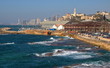 Zatoka Morza Śródziemnego w Tel Awiwie-Jaffie, promenada nadmorska w starej Jaffie, budynki, zaparkowane samochody, skały w wodzie, w tle, nieco rozmyte, drapacze chmur nowoczesnej częsci miasta