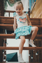 A Little Blond Girl In A White And Blue Dress In Polka Dots And Light Socks Descends A Wooden Ladder Crawling