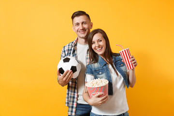 Wall Mural - Young fun couple, woman man, football fans holding soccer ball, bucket of popcorn, plastic glass of cola, cheer up support team, isolated on yellow background. Sport family leisure lifestyle concept.