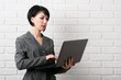 © soleg - business woman with laptop, dressed in a gray suit poses in front of a white wall