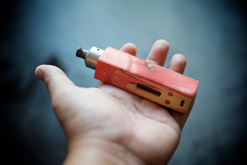 high end red natural stabilized wood box mods with rebuildable dripping atomizer vape gear in hand on dark grey texture background, vaporizer equipment, selective focus with shallow depth of field