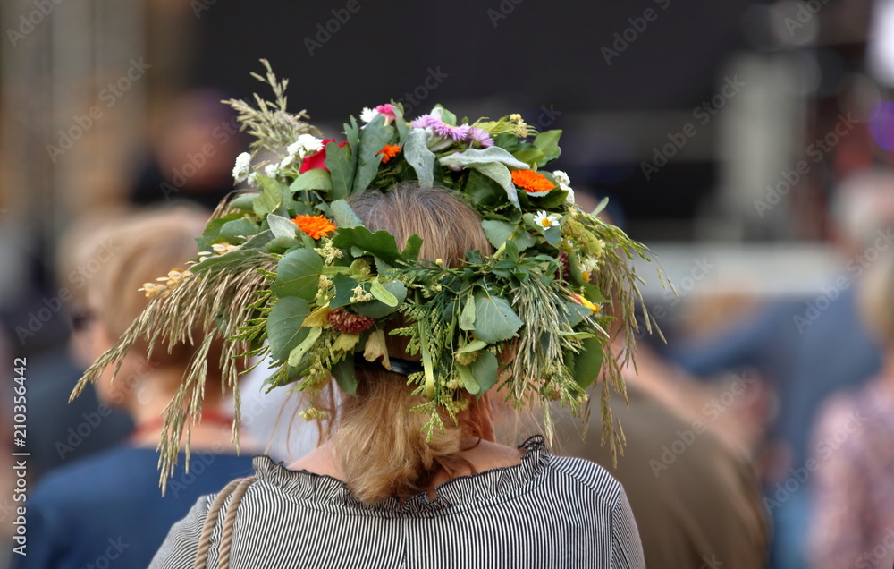 Kobieta, blond włosy, stoi tyłem (widoczna głowa i część ramion), w roślinnym, kwietnym, kolorowym wianku na głowie, w tle rozmyty tłum ludzi - obrazy, fototapety, plakaty 