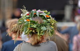 Fototapeta  - Kobieta, blond włosy, stoi tyłem (widoczna głowa i część ramion), w roślinnym, kwietnym, kolorowym wianku na głowie, w tle rozmyty tłum ludzi