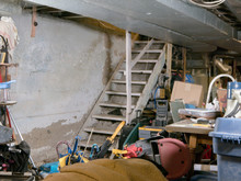 Concrete Wall Provides Text Space Inside Residential Unfinished Basement Being Used For Storage, Cluttered With Boxes, Bins, And Other Random Forgotten Items Of A Disposable Consumerist Lifestyle