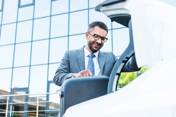 smiling businessman putting luggage into car on parking