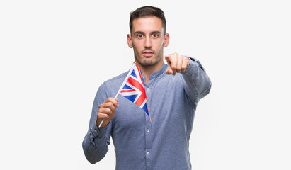 Handsome young man holding a flag of United Kingdom pointing with finger to the camera and to you, hand sign, positive and confident gesture from the front