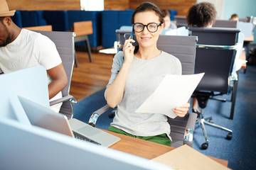 Wall Mural - Young smiling businesswoman in eyeglasses talking by mobile phone while sitting by workplace in open space