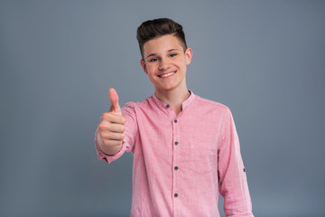 Pleased by everything. Upbeat teenage boy showing a thumbs up and grinning while posing isolated on a blue-grey background