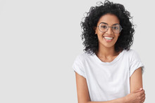 Waist Up Portrait Of Beautiful Young Dark Skinned Female With Positive Expression, Has Afro Hairdo, Feels Happy After Successful Bargain, Wears Big Spectacles, Isolated Over White Background