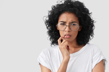 Wall Mural - Close up portrait of pretty serious displeased African American female frowns face, tries to concentrate and remeber something, wears round glasses and white t shirt, isolated on studio background
