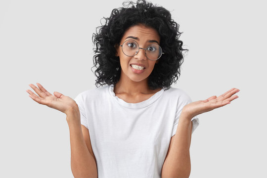 Clueless African American female with curly hair, looks doubtfully, clenches teeth and shrugs with shoulders, dressed in casual white t shirt, isolated over blank wall. People, hesitation concept