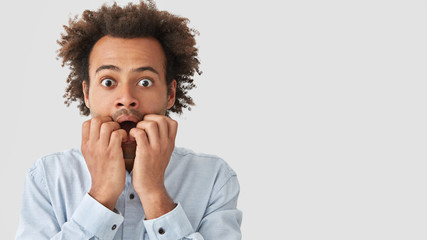 Wall Mural - Studio shot of embarrassed mixed race male with nervous expression, bites finger nails, worries before interview, has scared expression, Afro hairstyle, poses against white blank wall with free space