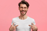 Fototapeta  - Horizontal shot of handsome cheerful hipster male indicates at white t shirt, shows blank space for your logo, being in high spirit, isolated over pink blank wall. Positive emotions and advertisement