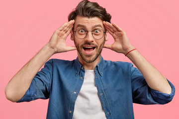 Wall Mural - Overjoyed surprised young Caucasian male with positive surprised expression, keeps hands on head, sees something unbelievable, wears spectacles and denim shirt, poses against pink blank wall