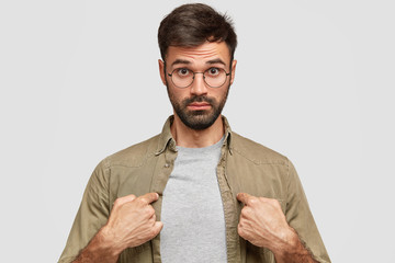 Wall Mural - Indoor shot of puzzled young Caucasian male looks with surprised expression while points at himself, dressed in shirt, has thick beard, wears round glasses, isolated over white studio background