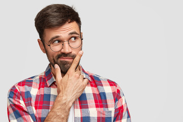 Thoughtful macho man holds chin and looks with hesitation aside, being unshaved, dressed in checkered shirt, stands against white wall with copy space for your advertisement. Pensive hipster