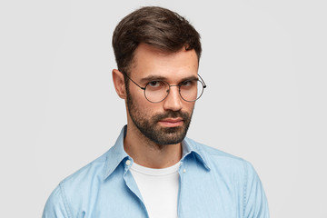 Wall Mural - Portrait of serious young male journalist wears round glasses and blue shirt, has confident look at camera, recieves important task from boss, listens attentively, isolated over white background.