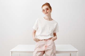 Wall Mural - Horizontal shot of attractive stylish and feminine female with natural ginger hair in bun hairstyle, leaning on table and holding hand in pockets, smiling broadly in sunglasses over grey wall