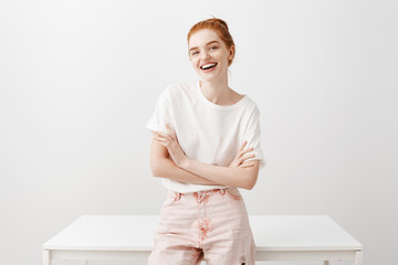 Wall Mural - I like what you say. Attractive joyful european female with ginger hair in stylish pink jeans, leaning on table and holding hands crossed on chest in confident pose, smiling broadly at camera