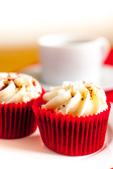 Two red velvet cupcakes with a white cup and saucer in the background