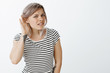 © Cookie Studio - Repeat louder, cannot hear your whisper. Portrait of intense confused attractive female student in striped t-shirt, bending towards camera, holding hand near ear, gossiping or eavesdropping