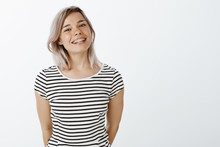 Studio Shot Of Cute Caucasian Girlfriend With Blond Hair And Braces On Teeth Standing In Casual Striped T-shirt And Smiling Broadly At Camera, Holding Hands Behind, Enjoying Great Sunny Day