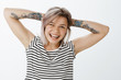 © Cookie Studio - Be happy and never give up. Carefree attractive european female with fair hair and tattoos, holding hands behind head, standing in relaxed pose and smiling broadly, showing shiny cute braces