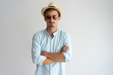 Serious handsome man in cool sunglasses crossing arms on chest and looking at camera. Confident young guy in straw hat wearing blue shirt. Summer time concept