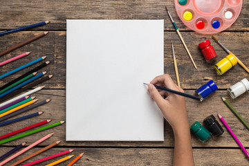 Top view of Girl hand drawing empty paper and colorful pencils, child painting using watercolors and paint brush on old wooden table