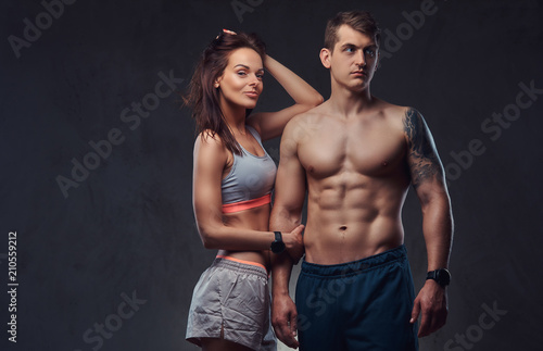Fotografía Attractive couple, a slim brunette female wearing sports bra and shorts and handsome shirtless guy cuddling in a studio on a dark textured background