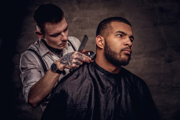 Old-fashioned professional tattooed hairdresser does a haircut to an African American client. Isolated on dark textured background.