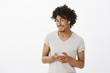 © Cookie Studio - Curious good-looking male with afro haircut, tilting head and smiling joyfully, holding smartphone and wearing wireless earphones, talking with friends via device, standing over grey wall