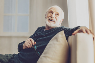 Pleasurable time. Joyful senior man smiling while enjoying his rest at home