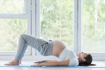 Beautiful young pregnant woman doing yoga or fitness exercise, lotus pose on the blue yoga mat at home. Happy and healthy pregnancy, meditation, calmness concept