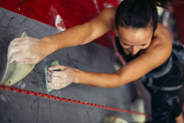 Wall Mural - Beautiful young strong woman climbing on rock artificial wall top view. Female climber insured on belaying harness by friends reaches top of the cliff indoors.