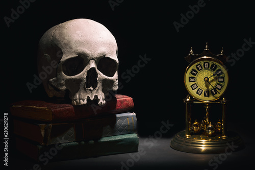 Fotografía Human skull on old books near retro vintage clock on black background under beam of light