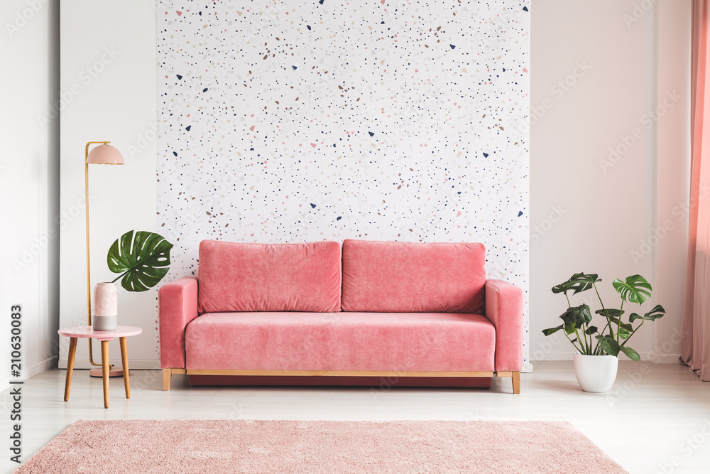 Pink couch between plant and lamp in bright living room interior with patterned wall. Real photo