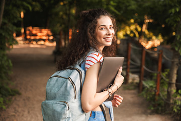 Sticker - Photo of caucasian young brunette woman 18-20 wearing backpack, walking through green park with silver laptop in hands