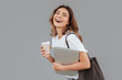 © Drobot Dean - Image of happy young woman 20s laughing and standing over gray wall isolated with silver laptop, and takeaway coffee in hands
