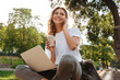 © Drobot Dean - Image of smiling modern woman sitting on bench in green park on summer day, and talking on smartphone while using silver laptop and drinking takeaway coffee