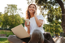 Image Of Smiling Modern Woman Sitting On Bench In Green Park On Summer Day, And Talking On Smartphone While Using Silver Laptop And Drinking Takeaway Coffee