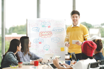 Young asian creative businessman standing and making presentation at modern office happy talking and brainstorming with teamwork behind transparent glass wall. Casual people business meeting concept.