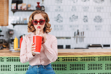 Beautiful lady in sunglasses and shirt sitting at the bar counter and drinking soda water in cafe