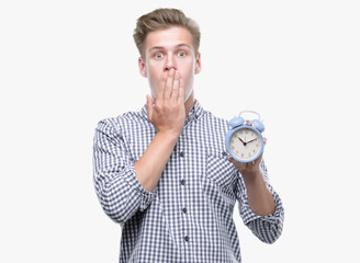 Wall Mural - Young handsome blond man holding alarm clock cover mouth with hand shocked with shame for mistake, expression of fear, scared in silence, secret concept