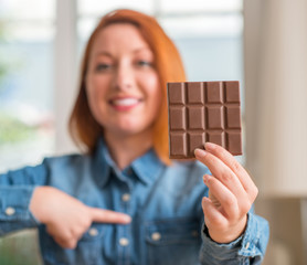 Wall Mural - Redhead woman holding chocolate bar at home with surprise face pointing finger to himself