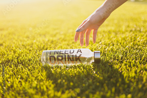 Cuadros en Lienzo Outstretched hand to bottle with fresh clean clear water with text inscription I need drink on green grass on sunshine lawn