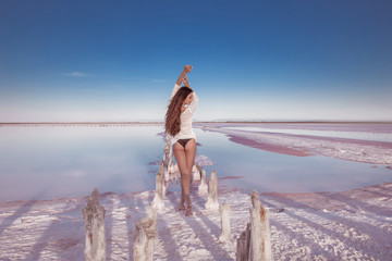 Wall Mural - Beautiful free sexy girl in white swimwear posing on salty beach at sunset. Sensual tanned woman with long healthy hair posing on salt pink lake enjoying nature landscape. Luxury summer vacation.