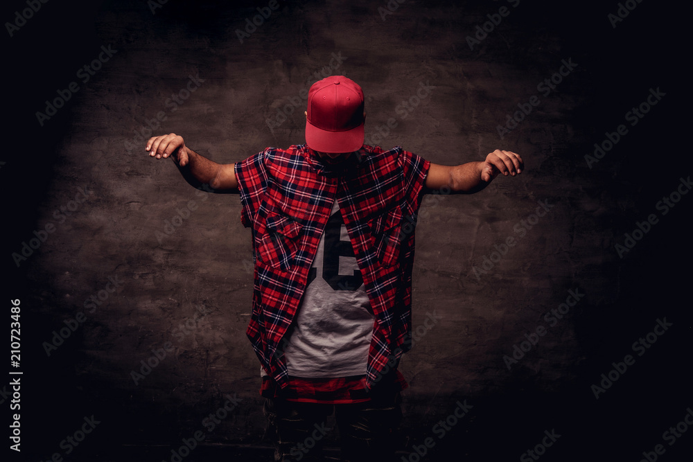 Portrait of an African-American dancer guy dressed in a red fleece shirt and cap at the studio. Isolated on dark textured background. - obrazy, fototapety, plakaty 