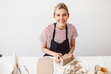 Cheerful girl in black apron and striped T-shirt sitting with piece of clay happily looking in camera at pottery studio